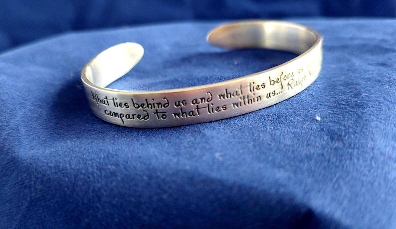 May include: A silver-colored cuff bracelet with engraved text. The inscription reads, "What lies behind us and what lies before us compared to what lies within us..." The bracelet is on a blue surface.