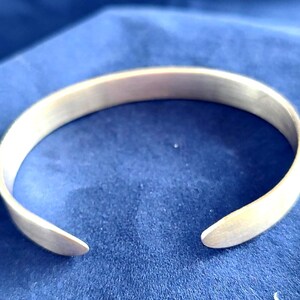 May include: A silver-colored cuff bracelet with a smooth, matte finish. The open-ended design allows for easy wear. The bracelet is displayed on a blue surface, highlighting its metallic sheen. A simple, elegant piece of jewelry.