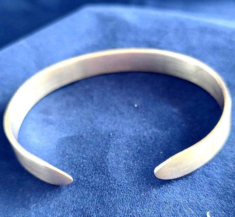 May include: A silver-colored cuff bracelet with a smooth, matte finish. The open-ended design allows for easy wear. The bracelet is displayed on a blue surface, highlighting its metallic sheen. A simple, elegant piece of jewelry.