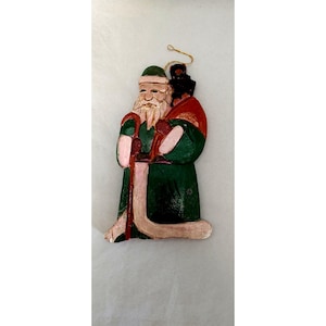 May include: A wooden Christmas ornament depicting a figure in a green robe with a white beard and a red and green hat. The ornament has a gold string for hanging and is painted with red and pink accents.