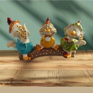 May include: A decorative figurine featuring three owl-like characters perched on a wooden branch. The figurines have white bodies, colorful outfits, and are adorned with small, sparkling embellishments. The background is a light teal color.