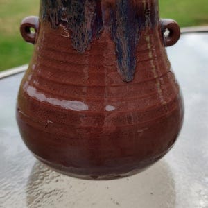 以下が含まれることがあります： 手作りのセラミックジャーと蓋。ジャーは濃い茶色で、表面はざらざらしており、2つの小さなハンドルが付いています。上部は濃い青と紫の釉薬が垂れています。蓋は濃いグレーです。