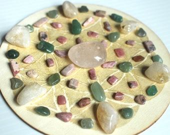 May include: A wooden circle with a white mandala design, decorated with various crystals and stones in shades of pink, green, and white.