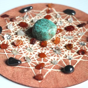 May include: A circular wooden board with a white geometric design. The board is decorated with various crystals, including clear quartz, carnelian, and black obsidian. A large turquoise stone is placed in the center of the board.
