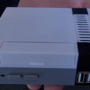 Op de afbeelding: Een witte en zwarte retro-videogames console met een slot voor een game cartridge, twee knoppen en een USB-poort.