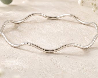 May include: A silver-toned bangle bracelet featuring a continuous wavy, undulating design. The metal surface has a subtle hammered texture, creating a reflective finish that catches the light along the curved edges of the circular jewelry piece.