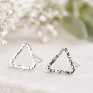May include: A pair of silver-toned stud earrings featuring an open triangle shape with a hammered, textured surface. Each earring has a post back and is crafted from a metallic material with a reflective finish, showcasing a geometric design.