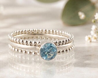 Könnte beinhalten: Ein Silberring mit einem zentralen, rund facettierten hellblauen Edelstein. Die Ringschiene besteht aus zwei äußeren Reihen kleiner Silberkügelchen und einer glatten mittleren Schiene, die den blauen Stein in einer Zargenfassung hält.