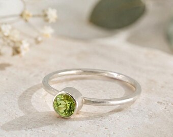 May include: A silver ring featuring a single, round, faceted lime-green peridot gemstone set in a simple bezel setting. The band is thin and polished, showcasing a smooth, metallic finish that complements the vibrant green stone.