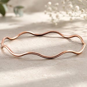 May include: A rose gold bangle bracelet featuring a continuous, smooth, wavy, or serpentine silhouette. The metal has a polished, reflective finish that catches the light, creating subtle highlights along the curved, undulating form of the circular band.
