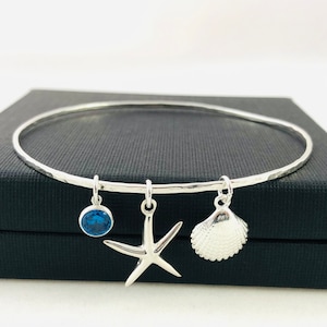 Könnte beinhalten: Ein silbernes Armreif-Armband mit drei Anhängern: einem blauen Edelstein, einem Seestern und einer Muschel.