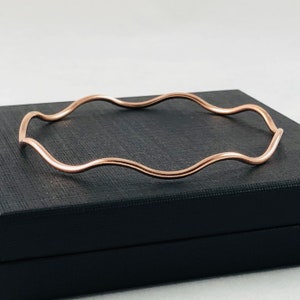 Op de afbeelding: Een roségouden armband met een golvend ontwerp. De armband is een doorlopende lus met een gladde, gepolijste afwerking. De armband wordt gepresenteerd op een zwart oppervlak.