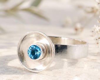 Puede incluir: Un anillo de plata con una banda ancha y plana y un engaste en forma de disco cóncavo. En el centro del disco hay una pequeña piedra preciosa azul redonda y facetada. El metal tiene un acabado pulido y el diseño está centrado.