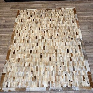 May include: Rectangular rug with a patchwork design. The rug features a pattern of rectangular pieces in shades of beige, cream, and brown. The edges of the rug are a darker brown. The rug is made of a natural material.