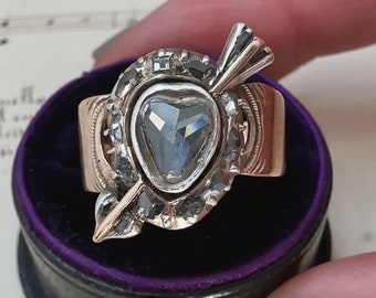 May include: A heart-shaped diamond ring with an arrow piercing through it. The ring is set in gold and has a silver band with black diamonds. The ring is in a black velvet ring box.