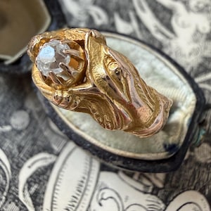 May include: A gold-toned ring with a large, clear gemstone. The ring's design features intricate, organic shapes that cradle the central stone. The ring is presented in a vintage, velvet-lined box, suggesting a piece of antique jewelry.