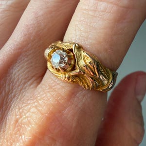 May include: A gold ring featuring a central, clear gemstone. The ring has an ornate, detailed design with a floral or leaf-like pattern. The ring is a warm gold color, and the gemstone is round and faceted.