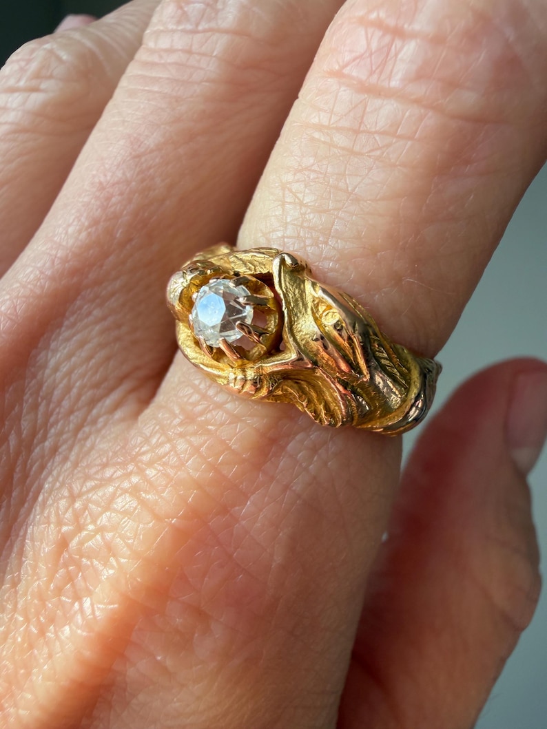 May include: A gold ring featuring a central, clear gemstone. The ring has an ornate, detailed design with a floral or leaf-like pattern. The ring is a warm gold color, and the gemstone is round and faceted.