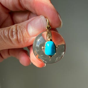 Op de afbeelding: Een heldere, ronde hanger met een gouden beugel en accenten. De hanger heeft een levendige turkooizen steen in het midden. De gouden zetting heeft ingewikkelde details. De hanger wordt door een hand omhoog gehouden.