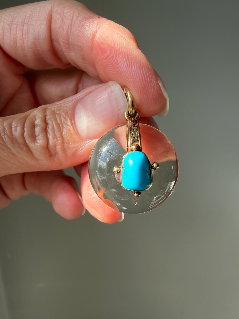 Op de afbeelding: Een heldere, ronde hanger met een gouden beugel en accenten. De hanger heeft een levendige turkooizen steen in het midden. De gouden zetting heeft ingewikkelde details. De hanger wordt door een hand omhoog gehouden.