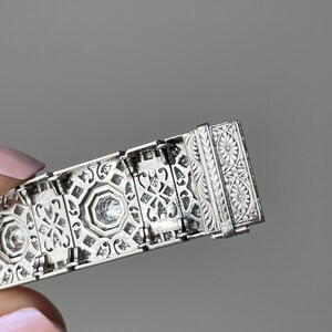 K&ouml;nnte beinhalten: Ein silbernes Armband mit komplizierten geometrischen Mustern und funkelnden Diamanten. Das Armband hat einen detaillierten Verschluss mit floralen Motiven.