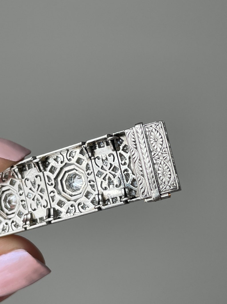K&ouml;nnte beinhalten: Ein silbernes Armband mit komplizierten geometrischen Mustern und funkelnden Diamanten. Das Armband hat einen detaillierten Verschluss mit floralen Motiven.