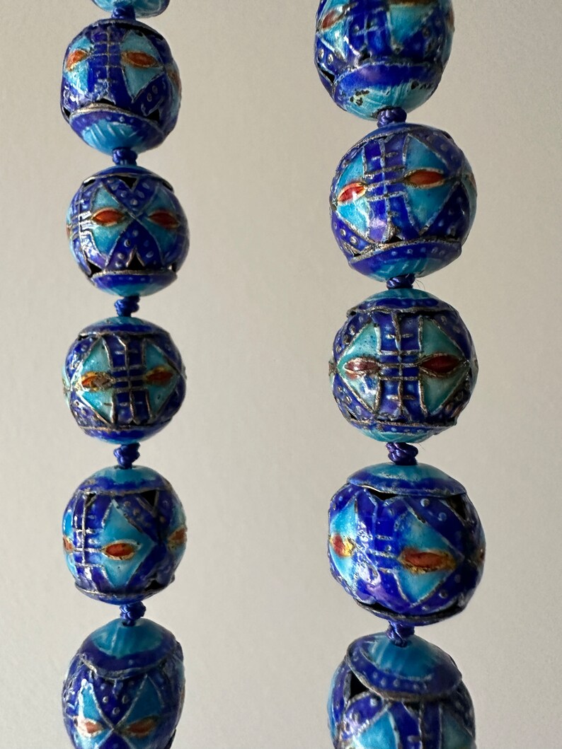 Puede incluir: Una larga hebra de cuentas de esmalte azul y blanco con intrincados patrones geom&eacute;tricos. Las cuentas est&aacute;n enhebradas en un cord&oacute;n delgado y tienen una base de metal plateado.