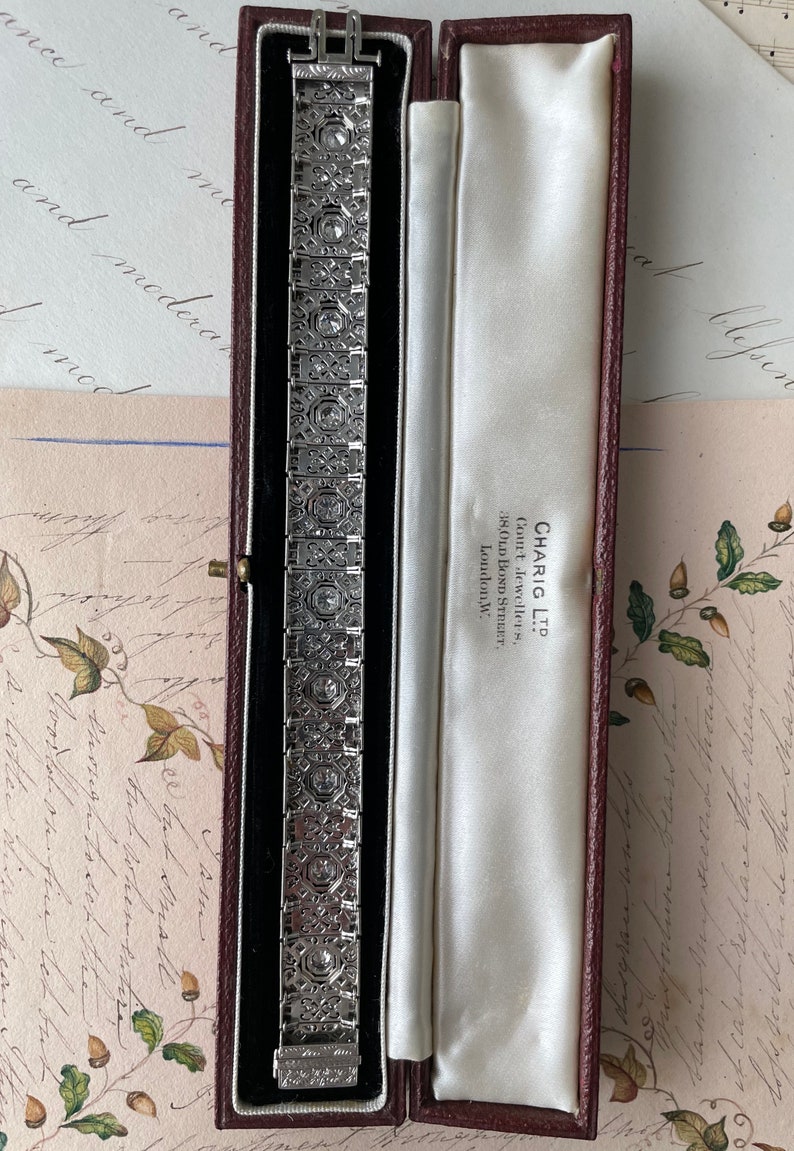K&ouml;nnte beinhalten: Ein silbernes Armband mit geometrischem Design und einer Reihe von Diamanten, die in der Mitte jedes Gliedes eingesetzt sind. Das Armband befindet sich in einer roten Samtbox mit wei&szlig;er Satin-Auskleidung. Die Box tr&auml;gt ein Etikett mit der Aufschrift "CHARIG LTD. Court Jewellers, 38, OLD BOND STREET. London, W."