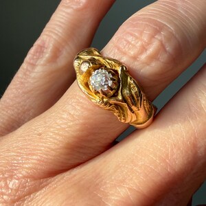 May include: A gold ring featuring a central, clear gemstone. The ring has an ornate, textured design with a floral or leaf-like motif. The ring is set against a blurred background, highlighting the jewelry's details.