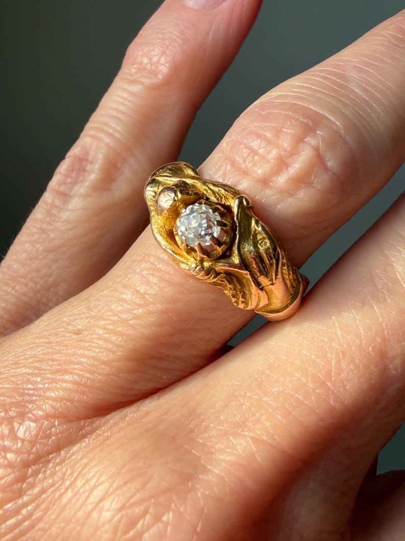 May include: A gold ring featuring a central, clear gemstone. The ring has an ornate, textured design with a floral or leaf-like motif. The ring is set against a blurred background, highlighting the jewelry's details.