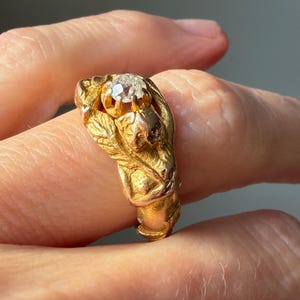 May include: A gold ring featuring a central, clear gemstone. The ring has an ornate design with sculpted details, including leaves and a small figure. The ring is held by a hand, showcasing its intricate craftsmanship and vintage style.