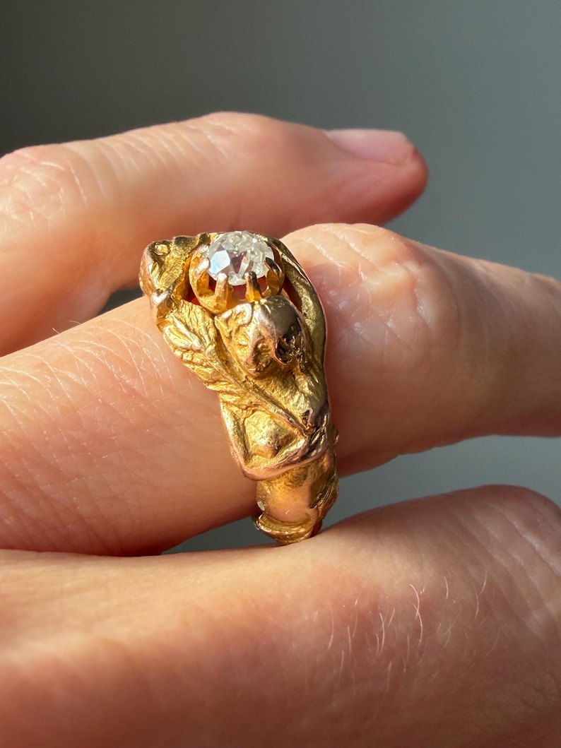 May include: A gold ring featuring a central, clear gemstone. The ring has an ornate design with sculpted details, including leaves and a small figure. The ring is held by a hand, showcasing its intricate craftsmanship and vintage style.