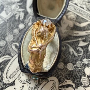 May include: A gold ring featuring a sculpted figure holding a clear gemstone. The ring is set in a black jewelry box with a light-colored interior. The ring's design includes floral and human-like elements.