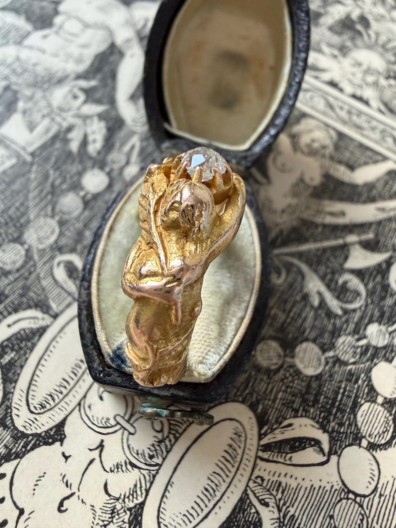 May include: A gold ring featuring a sculpted figure holding a clear gemstone. The ring is set in a black jewelry box with a light-colored interior. The ring's design includes floral and human-like elements.