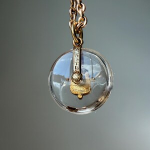 Op de afbeelding: Een heldere, ronde hanger met een goudkleurige metalen ketting. De hanger heeft een goudkleurig decoratief element aan de binnenkant. De ketting is gemaakt van in elkaar grijpende schakels. De achtergrond is effen grijs.