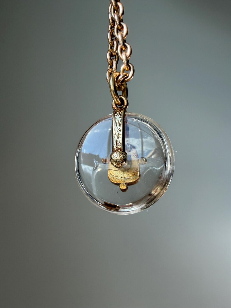 Op de afbeelding: Een heldere, ronde hanger met een goudkleurige metalen ketting. De hanger heeft een goudkleurig decoratief element aan de binnenkant. De ketting is gemaakt van in elkaar grijpende schakels. De achtergrond is effen grijs.