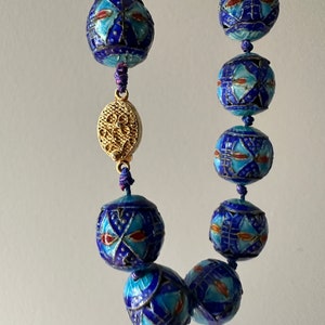 Puede incluir: Un collar azul y dorado con cuentas redondas. Las cuentas est&aacute;n decoradas con intrincados patrones en azul, blanco y rojo. El collar tiene un cierre dorado.