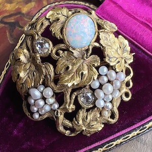 Puede incluir: Un broche de oro con una gran piedra de ópalo rodeada de un diseño de vid con detalles de perlas.
