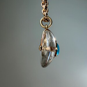 Op de afbeelding: Een goudkleurige ketting hangt een heldere, druppelvormige hanger met een blauwe edelsteen op. De hanger is facetgeslepen en vangt het licht. De sieraden zijn geplaatst tegen een zachte grijze achtergrond.