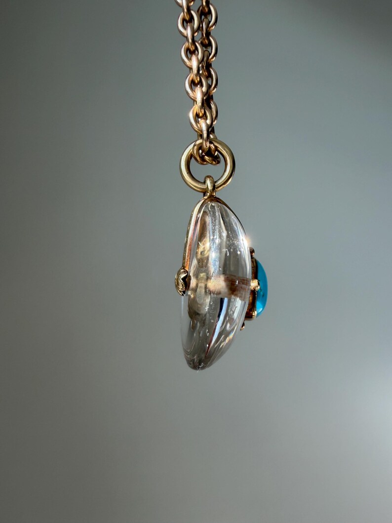 Op de afbeelding: Een goudkleurige ketting hangt een heldere, druppelvormige hanger met een blauwe edelsteen op. De hanger is facetgeslepen en vangt het licht. De sieraden zijn geplaatst tegen een zachte grijze achtergrond.