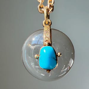 Op de afbeelding: Een goudkleurige ketting met een transparante, ronde hanger. De hanger heeft een heldere turkooizen steen in goud gezet. De ketting en verbindingsstukken zijn ook goudkleurig. De hanger heeft een diameter van ongeveer 3 cm.