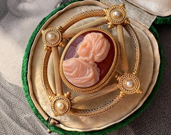 Puede incluir: Un broche de camafeo dorado con un retrato de camafeo blanco de una mujer con turbante. El broche está engastado en un marco geométrico con perlas y se encuentra en una caja de terciopelo verde.