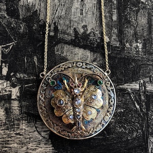 May include: A gold pendant necklace with a round, ornate, enamel-painted butterfly design. The butterfly has a detailed body and wings with white diamonds and green and red gemstones. The pendant is suspended from a gold chain.