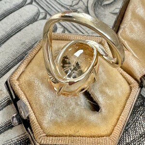 May include: A gold ring with a large, oval-shaped, light brown gemstone. The ring is set in a twisted band and is sitting in a small, velvet-lined box.
