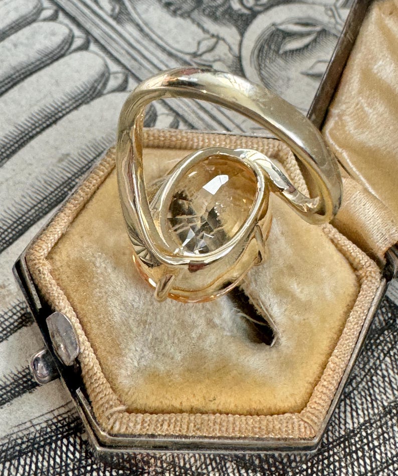 May include: A gold ring with a large, oval-shaped, light brown gemstone. The ring is set in a twisted band and is sitting in a small, velvet-lined box.