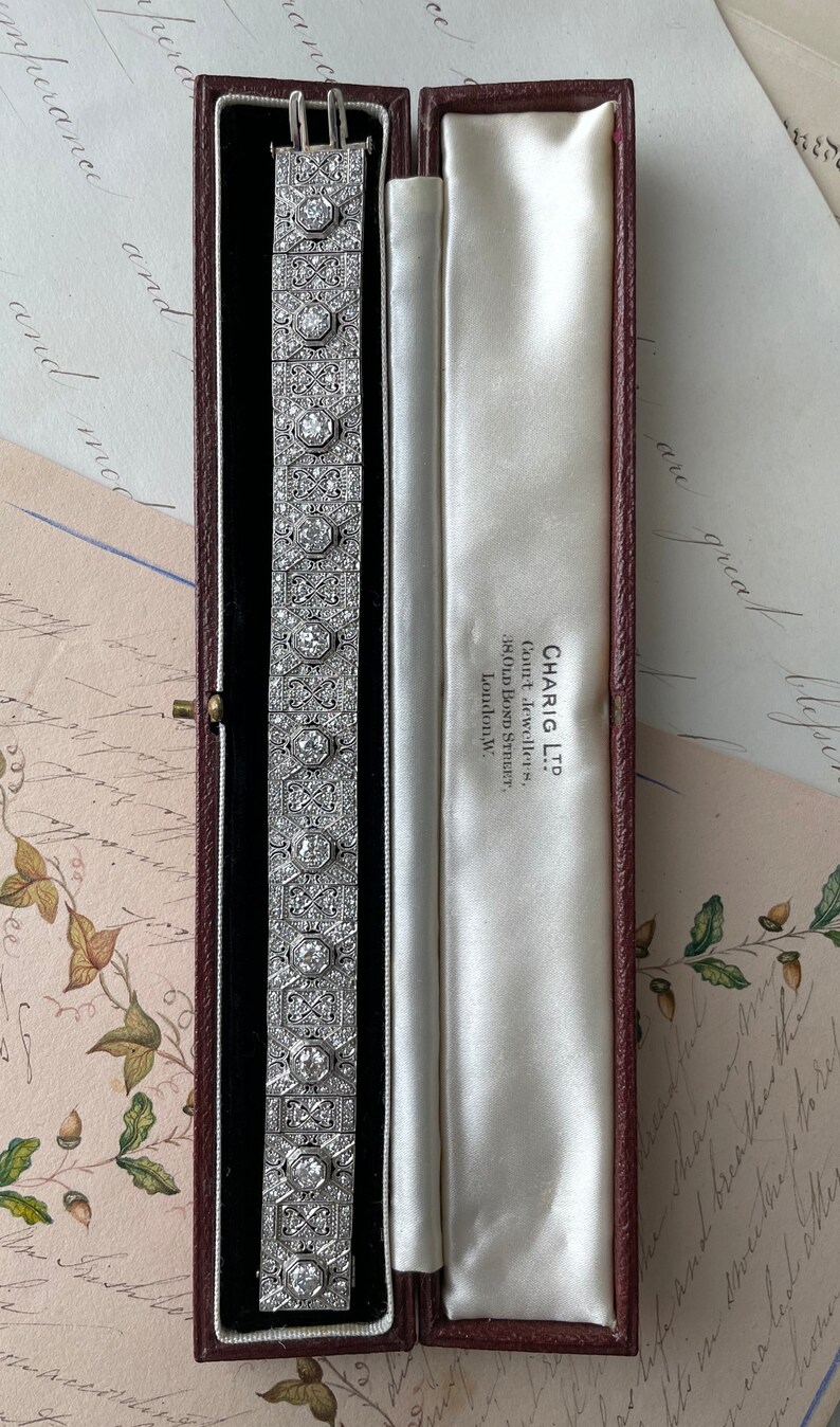 K&ouml;nnte beinhalten: Ein silbernes Armband mit geometrischem Design, besetzt mit Diamanten. Das Armband befindet sich in einer roten Samtbox mit wei&szlig;er Satin-Auskleidung. Die Box tr&auml;gt ein Etikett mit der Aufschrift "CHARIG LTD.  Con't Jewellers, 38,OLD BOND STREET, London,W."