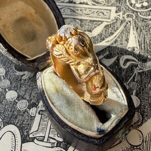 May include: A gold ring with a detailed design, featuring a central clear stone and ornate embellishments. The ring is displayed in an open, vintage-style black jewelry box with a light blue interior. The background has a black and white patterned design.