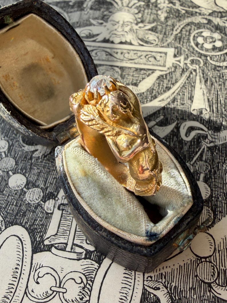 May include: A gold ring with a detailed design, featuring a central clear stone and ornate embellishments. The ring is displayed in an open, vintage-style black jewelry box with a light blue interior. The background has a black and white patterned design.