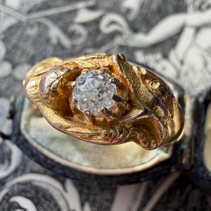 May include: A gold ring with a large, clear diamond center stone. The ring has an ornate, textured design around the diamond, with a vintage aesthetic. The ring is set against a patterned background.