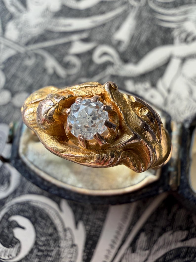 May include: A gold ring with a large, clear diamond center stone. The ring has an ornate, textured design around the diamond, with a vintage aesthetic. The ring is set against a patterned background.
